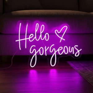 Può includere: Un'insegna al neon rosa acceso mostra le parole "Hello gorgeous" in corsivo con un cuore. L'insegna è posizionata davanti a un divano, creando un'atmosfera calda.