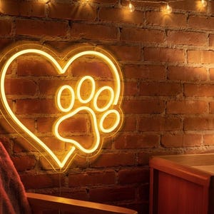 Pode incluir: Uma placa de néon em forma de coração com uma pegada de pata no interior, iluminada com um brilho amarelo quente. Montada numa parede de tijolo vermelho, criando um ambiente acolhedor. Luzes decorativas.