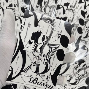 May include: Black and white sticker featuring a musician playing a double bass. The illustration has a vintage style with the word "Bassy" in a decorative font. The background shows other stickers with the same design.