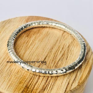 Puede incluir: Un brazalete de metal plateado martillado. El brazalete tiene una superficie texturizada y se muestra sobre una superficie de madera. El texto "madebycoppersring" es visible en la superficie de madera.
