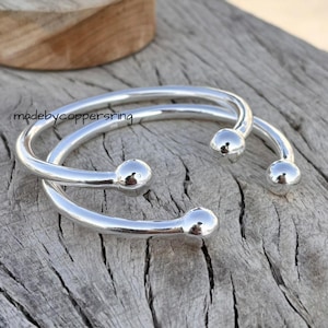 Könnte beinhalten: Zwei silberfarbene, offene Armreifen mit abgerundeten Kugelenden. Die Armreifen sind gebogen und liegen auf einer verwitterten, grauen Holzoberfläche. Der Text "madebycoppersring" ist sichtbar.