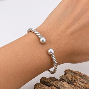 Può includere: Un bracciale a fascia color argento caratterizzato da una texture intrecciata simile a una corda lungo il corpo e due estremità sferiche lisce e lucide. Il design aperto consente una vestibilità flessibile con finitura metallica.