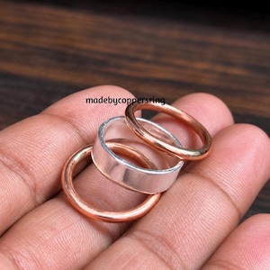 Könnte beinhalten: Ein Set aus drei Ringen, bestehend aus zwei dünnen Kupferringen und einem breiteren silberfarbenen Ring. Die Ringe sind ineinandergestapelt. Der Text "madebycoppersring" ist darüber zu sehen. Die Kupferringe haben eine glatte, metallische Oberfläche.
