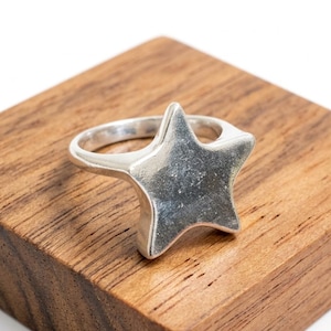 Könnte beinhalten: Ein silberfarbener Ring mit einem markanten fünfzackigen Sternmotiv auf der Vorderseite. Der Stern hat eine glatte, leicht texturierte Oberfläche mit metallischem Glanz. Die Ringschiene ist abgerundet und mit der Rückseite des Sterns verbunden.