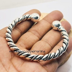 Könnte beinhalten: Ein silberfarbenes Armband mit einem gedrehten Seildesign. Das Armband hat abgerundete Endkappen und wird in einer Hand gehalten. Der Text "madebycoppersring" ist auf dem Armband zu sehen.