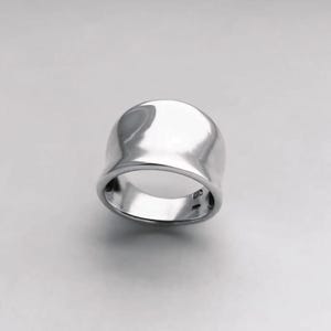 Puede incluir: Un anillo de plata pulida con una parte superior redondeada y lisa y una banda gruesa. El anillo tiene una superficie reflectante, que capta la luz. El diseño es simple y moderno, adecuado para el uso diario.