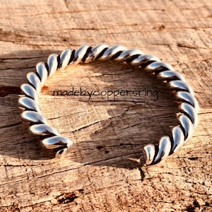 Könnte beinhalten: Ein silberfarbenes Armreif-Armband mit einem gedrehten Seildesign. Das offene Armband liegt auf einer Holzoberfläche. Der Text "madebycoppersring" ist sichtbar. Das Design des Armbands erinnert an ein Seil.