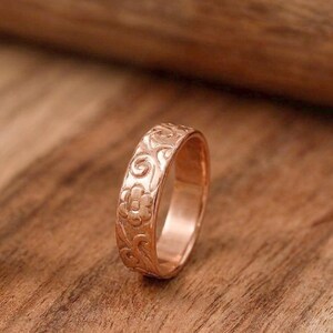 Könnte beinhalten: Ein Ring aus Roségold mit einem geprägten Blumen- und Rankenmuster auf der Außenseite. Das Metall hat eine polierte Oberfläche, die das Licht auf den detaillierten Schnitzereien der Blumen und geschwungenen Rankenmotive reflektiert.