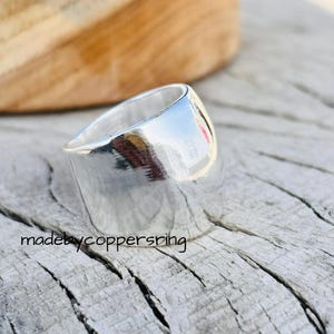 Könnte beinhalten: Ein breiter, polierter Silberring mit glatter, reflektierender Oberfläche. Der Ring hat ein leicht abgewinkeltes Design und ist vor einem rustikalen Holzhintergrund platziert. Der Text "madebycoppersring" ist sichtbar.