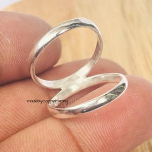 May include: A silver ring with a unique, open design. The ring features two bands that curve and overlap, creating a stylish and modern look. The text "madebycoppersring" is visible on the ring.