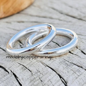 Könnte beinhalten: Zwei glänzende, silberne Ringe mit glattem, abgerundetem Design. Die Ringe werden auf einer verwitterten, grauen Holzoberfläche gezeigt. Der Text "madebycoppersring" ist sichtbar.
