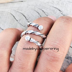 Könnte beinhalten: Ein silberfarbener Spiralring mit einem gedrehten Seildesign und einer kleinen Kugel an jedem Ende. Der Ring ist um einen Finger gewickelt. Der Text "madebycoppersring" ist sichtbar.