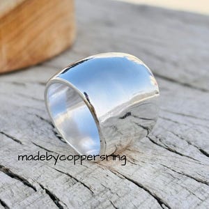 Puede incluir: Un anillo ancho de plata pulida con una superficie lisa y reflectante. El anillo está sobre un fondo de madera envejecida. El texto "madebycoppersring" es visible en la madera.