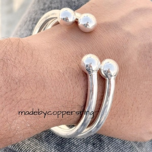 May include: A silver cuff bracelet with a distinctive design. The bracelet has three parallel bands that curve around the wrist, with two spherical accents on top. The text "madebycoppersring" is visible on the arm.
