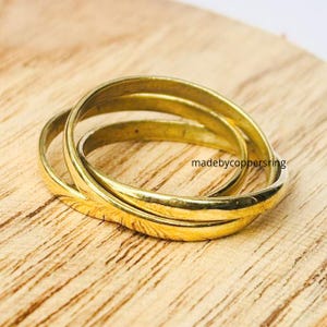 Könnte beinhalten: Drei ineinandergreifende, goldfarbene Ringe. Die Ringe sind glatt und glänzend, mit einem warmen, gelben Farbton. Die Ringe liegen auf einer hellbraunen Holzoberfläche. Der Text "madebycoppersring" ist sichtbar.