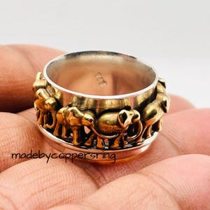 Könnte beinhalten: Ein Silberring mit einem goldfarbenen Band mit einem sich wiederholenden Muster von Elefanten. Der Ring hat ein rotierendes Mittelband und der Text "madebycoppersring" ist auf der Seite sichtbar.