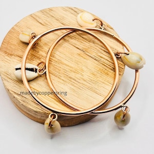 May include: Two copper-colored hoop earrings with dangling cowrie shells. The earrings are resting on a round wooden surface. The text "madebycoppersring" is visible on the wooden surface.