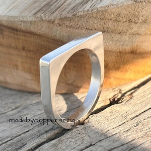Könnte beinhalten: Ein silberfarbener Ring mit einem geometrischen, quadratisch geformten Oberteil und einem abgerundeten Innenband. Der Ring ist vor einem Holzgrund platziert. Der Text "madebycoppersring" ist sichtbar.