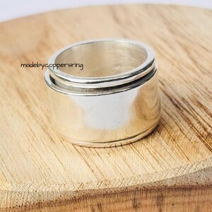 Könnte beinhalten: Ein silberfarbener Ring mit breitem Band und strukturiertem Design. Der Ring hat eine zylindrische Form mit einem drehbaren Außenband. Er wird auf einer Holzoberfläche präsentiert. Der Text "madebycoppersring" ist sichtbar.