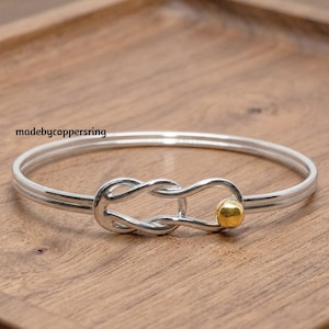 Może przedstawiać: Srebrna bransoletka typu bangle z centralnym węzłem w stylu nieskończoności i małym złotym akcentem w kształcie kulki. Napis madebycoppersring jest widoczny po lewej stronie bransoletki. Bransoletka ma gładkie, polerowane wykończenie.