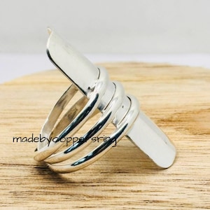 Pode incluir: Um anel de prata com um design único. O anel apresenta uma série de faixas curvas que envolvem o dedo, com um elemento retangular plano no topo. O texto "madebycoppersring" é visível na superfície de madeira.