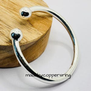 Könnte beinhalten: Ein silberfarbenes Armreif-Armband mit einem abgerundeten, offenen Design. Jedes Ende des Armbands hat einen kleinen, kugelförmigen Akzent. Das Armband wird auf einer Holzoberfläche präsentiert. Der Text "madebycoppersring" ist sichtbar.