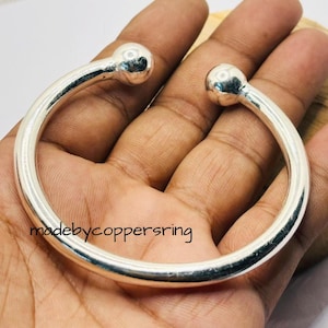 May include: A silver cuff bracelet with a rounded design. The bracelet has a smooth, polished finish and features a ball-shaped detail at each end. The text "madebycoppersring" is visible on the bracelet.