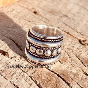 Può includere: Un anello d'argento con un design a strati. L'anello presenta una texture a forma di corda, una fila di piccole perle rotonde e una superficie liscia e lucida. L'anello è esposto su una superficie di legno. Il testo "madebycopperstring" è visibile.