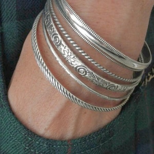 Könnte beinhalten: Ein Set aus fünf silberfarbenen Metallarmreifen, die zusammen getragen werden. Die Kollektion umfasst ein gedrehtes Seildesign, ein glattes Band, ein dünnes schlichtes Band, ein Band mit Blumen- und Wirbelgravuren sowie ein breiteres Band.