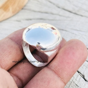 Könnte beinhalten: Ein polierter Silberring mit einem großen, abgerundeten Oberteil. Das Design des Rings weist eine glatte, reflektierende Oberfläche auf, die das Umgebungslicht einfängt. Der Ring wird in einer Hand gehalten, und der Text "madebycoppersring" ist sichtbar.