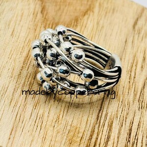 Könnte beinhalten: Ein silberner Ring mit mehreren Bändern und kleinen, runden Perlen, die sich oben gruppieren. Der Ring liegt auf einer Holzoberfläche, mit dem Text "madebycoppering" sichtbar.