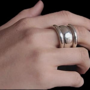 Könnte beinhalten: Ein silberner Ring mit einem einzigartigen Design. Der Ring hat ein zentrales, abgerundetes Band, das von zwei gebogenen, parallelen Bändern flankiert wird. Das polierte Metall reflektiert das Licht und erzeugt Glanzlichter und Schatten. Der Ring wird an einem Finger vor dunklem Hintergrund getragen.