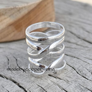 Könnte beinhalten: Ein Stapel silberner Ringe mit einem verschlungenen Design. Die Ringe bestehen aus einem glänzenden, metallischen Material und sind übereinander gestapelt. Die Ringe befinden sich auf einer Holzoberfläche. Der Text "madebycoppersring" ist sichtbar.