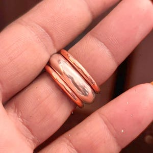 Könnte beinhalten: Ein Set aus drei gestapelten Ringen mit poliertem kupferfarbenem Finish. Die Ringe bestehen aus zwei schmaleren Bändern an den Außenkanten und einem breiteren, glatten, abgerundeten Band in der Mitte, die alle eine reflektierende Metalloberfläche aufweisen.