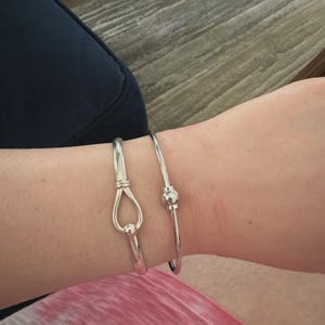 May include: Two silver-toned metal bangles worn on the wrist. One bangle features a teardrop-shaped loop design with a small sphere at the base. The second bangle is a thin, smooth wire band accented with a single small, polished spherical bead.