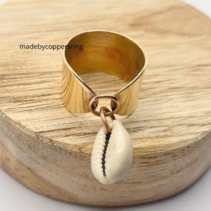 May include: A wide, gold-colored ring with a textured surface and a small, dangling cowrie shell. The ring is attached to a copper-colored loop. The text "madebycoppersring" is visible on the wooden surface.