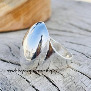 Puede incluir: Un anillo de plata pulida con una parte superior ovalada grande y alargada. El anillo refleja el entorno, creando un efecto de espejo. El anillo está sobre una superficie de madera envejecida. El texto "madebycoppersring" es visible.