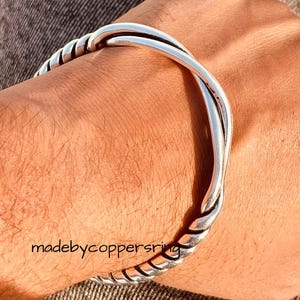 May include: A silver-colored cuff bracelet with a twisted rope design. The bracelet is worn on a wrist, with the text "madebycoppersring" visible. The bracelet has a smooth, polished finish.