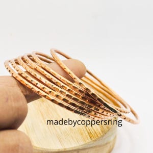 Könnte beinhalten: Eine Reihe gehammerter Kupferarmreifen. Die Armreifen haben eine strukturierte Oberfläche und eine warme, rötlich-braune Farbe. Der Text "madebycoppersring" ist auf der Holzoberfläche darunter zu sehen.