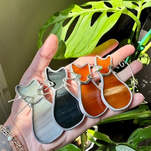 May include: Four stained glass cat ornaments in shades of white, gray, black, and orange. Each cat has a silver-colored metal outline and a wire loop for hanging. The ornaments are held in a hand, with green foliage in the background.