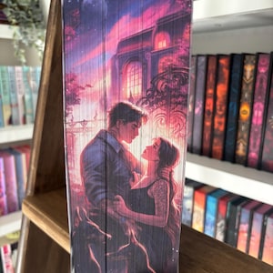 May include: A book with a romantic cover illustration. The artwork depicts a couple embracing in front of a building with a large arched window. The background features a sunset with shades of pink, purple, and orange. The book is displayed on a wooden shelf with other books.