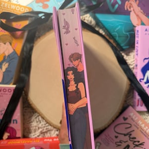 May include: A book with a pink spine featuring illustrations of a couple embracing, hearts, a microscope, and DNA strands. Other books with titles like "Deep End" and "Check & Mate" are visible.