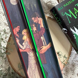 Może przedstawiać: Trzy książki z ilustrowanymi okładkami przedstawiającymi romantyczne sceny. Grzbiety są czerwone, zielone i czarne. Książka po prawej stronie ma tytuł "MATE" i nazwisko autora, Ali Hazelwood. Okładki przedstawiają pary i leśne krajobrazy.