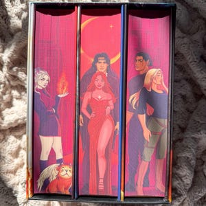 Könnte beinhalten: Ein Buchset mit drei Fantasy-Romanen mit lebendigen Cover-Illustrationen. Die Cover zeigen Charaktere in verschiedenen Posen, mit einem roten und rosa Farbschema. Das Set wird in einer dekorativen Box präsentiert.