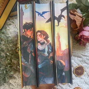 May include: Three hardcover books with matching covers. The covers depict a man and woman with dragons in the sky, set against a sunset backdrop. The book spines are black with gold detailing.