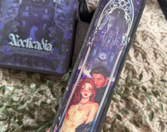 May include: A book with a purple and black cover featuring the title "Nocticadia" and an illustration of a gothic cathedral. The spine displays a romantic illustration of a couple. The book is held with a green sleeve.