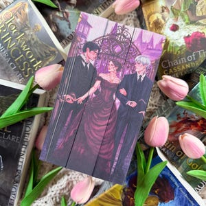 Op de afbeelding: Een verzameling fantasyromans met ge&iuml;llustreerde covers in tinten paars, zwart en wit. De covers tonen personages en sc&egrave;nes. Roze tulpen en groene bladeren zijn rond de boeken gerangschikt.