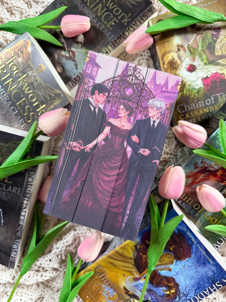 Op de afbeelding: Een verzameling fantasyromans met ge&iuml;llustreerde covers in tinten paars, zwart en wit. De covers tonen personages en sc&egrave;nes. Roze tulpen en groene bladeren zijn rond de boeken gerangschikt.