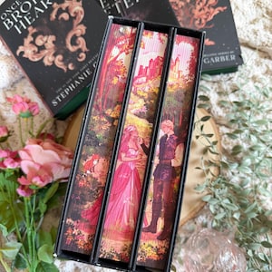 May include: A set of three hardcover books with dark covers and gold lettering, alongside a decorative box with a fantasy illustration. The books are titled "Once Upon a Broken Heart", "Ballad of Never After", and "Curse for True Love". The box features a forest scene with characters.
