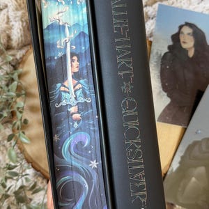 May include: A hardcover book with a black spine featuring the title "CALLIE HART - QUICKSILVER" in metallic lettering. The book's edge has a detailed illustration of a woman holding a sword, with a blue and purple color scheme.